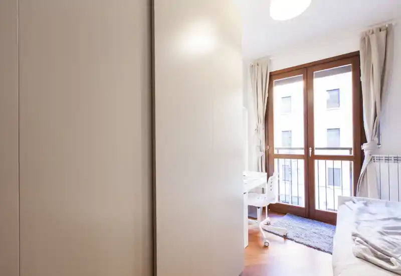 Bright, cozy bedroom with large floor-to-ceiling window, light curtains and a compact desk; neat bedding and a rug create a welcoming atmosphere.