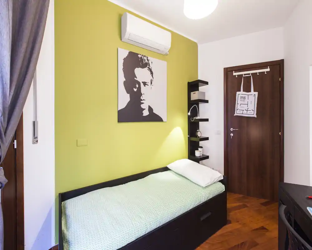 Piccola camera singola con letto ben rifatto contro una parete verde chiaro, condizionatore a muro, mensole a giorno e pavimento in legno caldo. Spazio luminoso, ordinato e ben arredato, ideale per una persona.