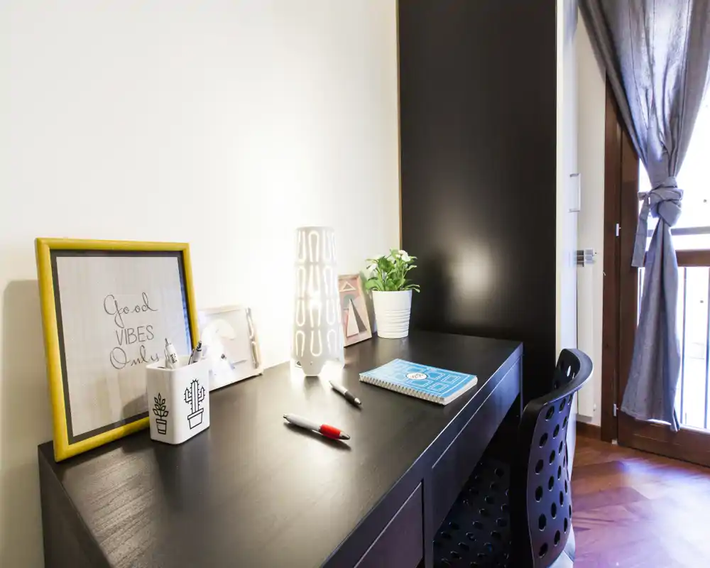 Angolo scrivania in camera ben illuminato con scrittoio in legno scuro, lampada decorativa, pianta e porta finestra con luce naturale.