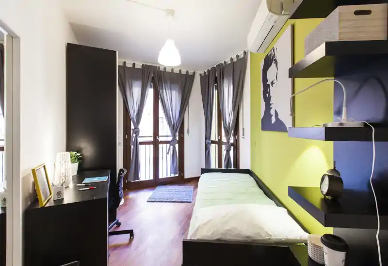 Camera singola moderna e luminosa con letto ordinato, scrivania e ampie finestre che lasciano entrare luce naturale. Spazio compatto ma ben arredato, ideale per uno studente o una persona sola.