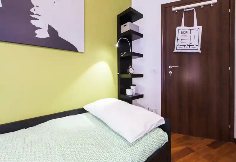 Camera da letto piccola e luminosa con letto singolo con copriletto a motivo, lampada da lettura, mensole verticali e porta in legno; quadro appeso che completa l'ambiente accogliente.
