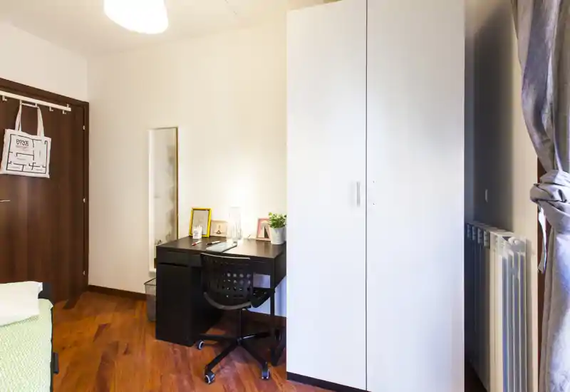 Piccolo angolo camera da letto ordinato con scrivania, armadio e letto parzialmente visibile sul lato sinistro. Pavimenti in legno caldo, sedia da studio e lampada creano un ambiente accogliente e ben illuminato, ideale per una persona sola.