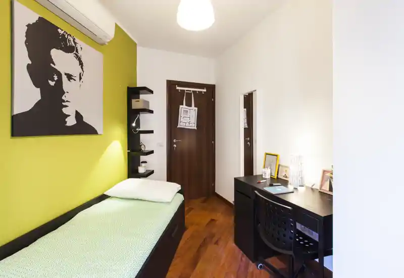 Camera singola compatta con letto singolo contro una parete verde, scrivania e sedia per lo studio, pavimento in legno caldo e quadro decorativo — luminosa e ben allestita per un annuncio.