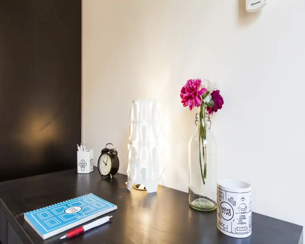 Un piano comodino ordinato con una lampada moderna, fiori freschi in una bottiglia, una sveglia e un taccuino — scatto luminoso e curato, perfetto per valorizzare i dettagli della stanza.