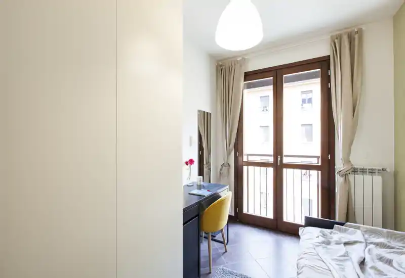 Camera da letto luminosa e moderna con una grande finestra/porta che fa entrare molta luce naturale, letto parzialmente visibile e una scrivania ordinata con sedia gialla — ideale per soggiorni confortevoli o smart working.