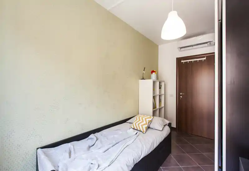 Camera singola compatta e luminosa con letto ben sistemato, cuscino a motivo chevron e scaffale accanto alla porta; ideale per uno studente o una persona singola.