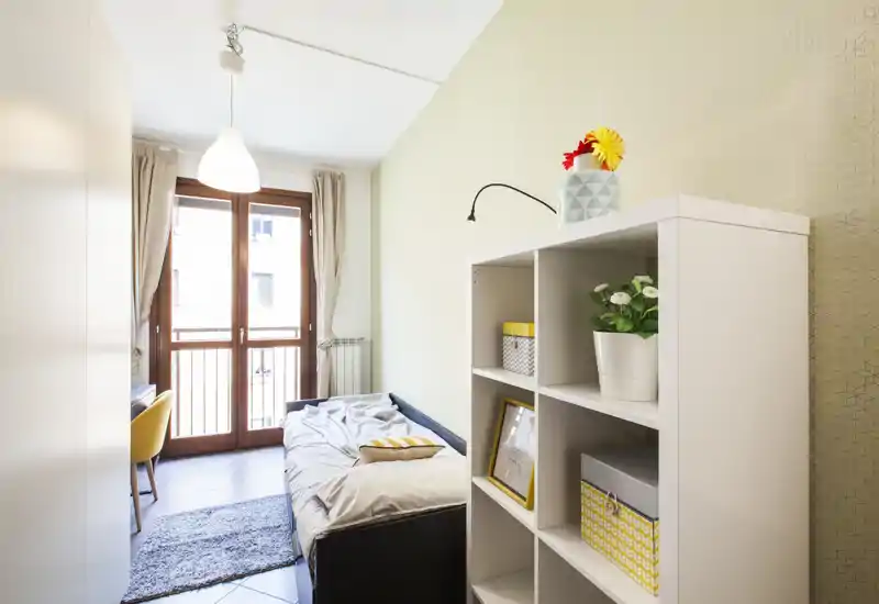 Camera da letto compatta e luminosa con letto singolo, libreria a cubi e porta finestra verso il balcone che lascia entrare molta luce naturale. Toni neutri, tessili morbidi e piante decorative rendono l’ambiente fresco e accogliente, perfetto per un annuncio.
