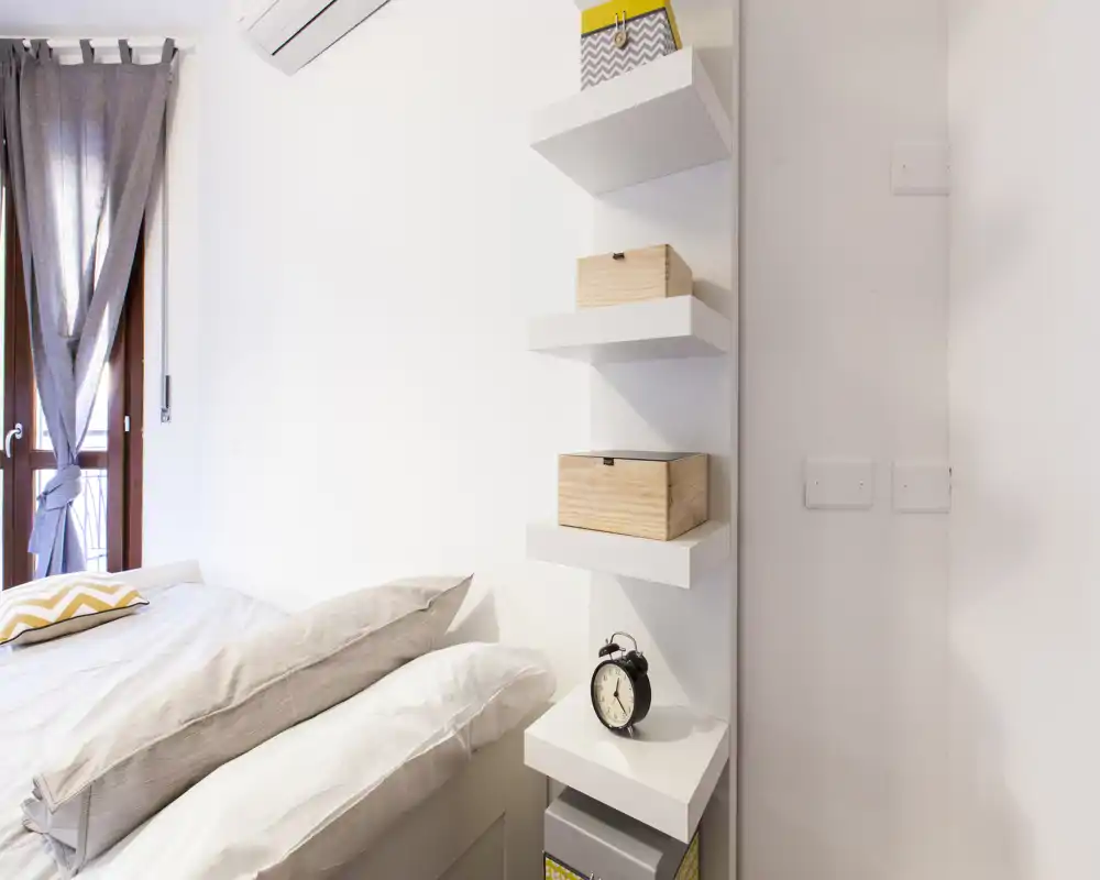 Rincón de dormitorio luminoso y moderno con cama bien hecha, cojines decorativos y estanterías de pared con cajas de almacenaje — perfecto para un alquiler compacto y con estilo.