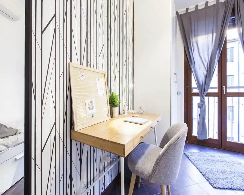 Rincón de dormitorio luminoso con escritorio de madera compacto y silla tapizada junto a la ventana/balcón; se aprecia una parte de la cama en el lateral izquierdo. Perfecto para una persona o como zona de estudio con luz natural.