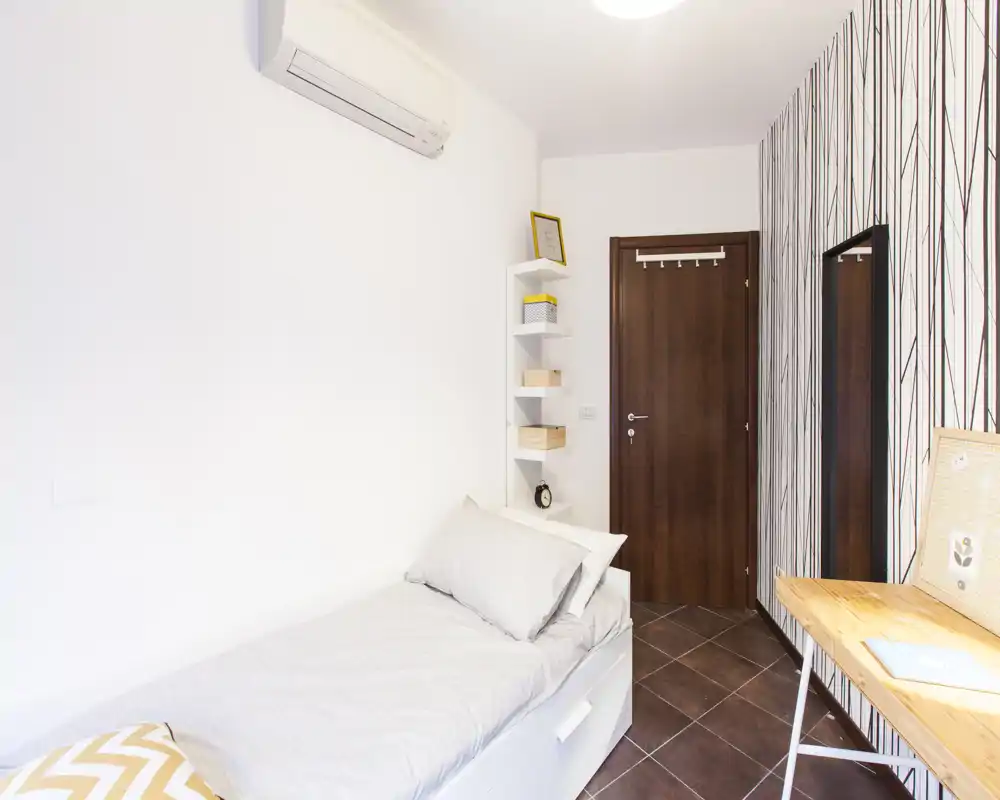 Dormitorio individual compacto con cama bien hecha, paredes blancas, aire acondicionado en la pared y un pequeño escritorio de madera junto a la pared con estampado — luminoso y funcional para una persona.