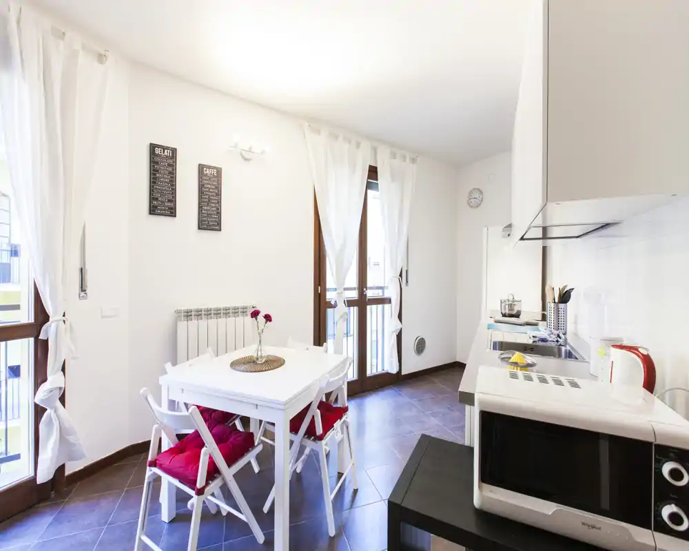 Cucina moderna e luminosa con un piccolo tavolo da pranzo. Pensili bianchi, un microonde sul piano e cuscini rossi rendono l'ambiente fresco e accogliente, perfetto per affitti brevi.