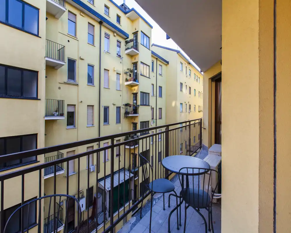 Balcone cittadino soleggiato con ringhiera in metallo e un piccolo tavolo rotondo con due sedie, affacciato su facciate gialle di condomini vicini — ideale per il caffè mattutino.