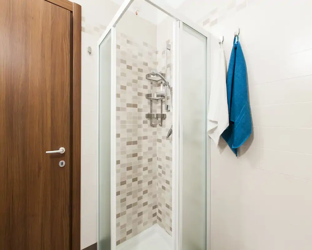 Bagno moderno compatto con box doccia ad angolo con porte in vetro satinato e piastrelle mosaico beige; due asciugamani appesi e porta in legno visibile.