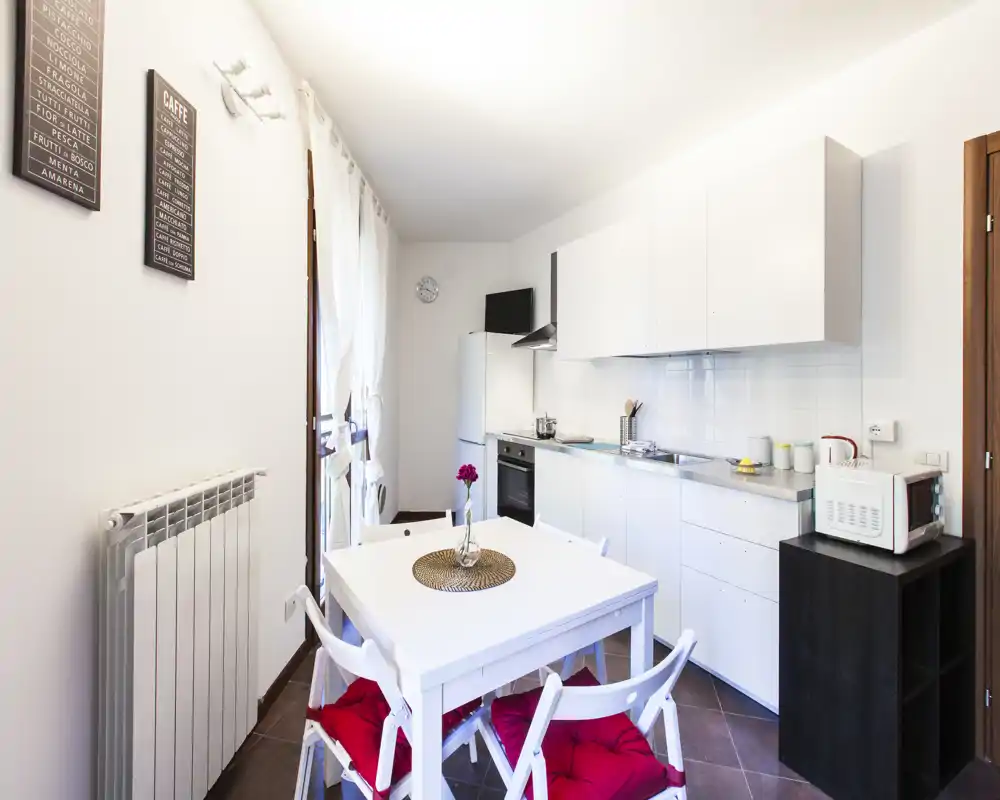 Angolo cottura moderno e luminoso con mobili bianchi, forno e frigorifero integrati e un tavolo da pranzo con cuscini rossi — perfetto per un appartamento compatto.