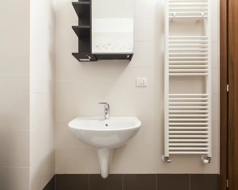 Area bagno compatta e pulita con lavabo a parete, pensile con specchio e scaldasalviette verticale — piastrelle chiare e finiture moderne.