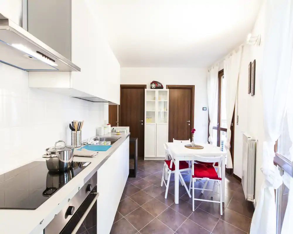 Cucina moderna e luminosa con pensili bianchi, piano cottura a induzione e un piccolo tavolo da pranzo con sedie dotate di cuscini rossi — perfetta per appartamenti compatti con tanta luce naturale.