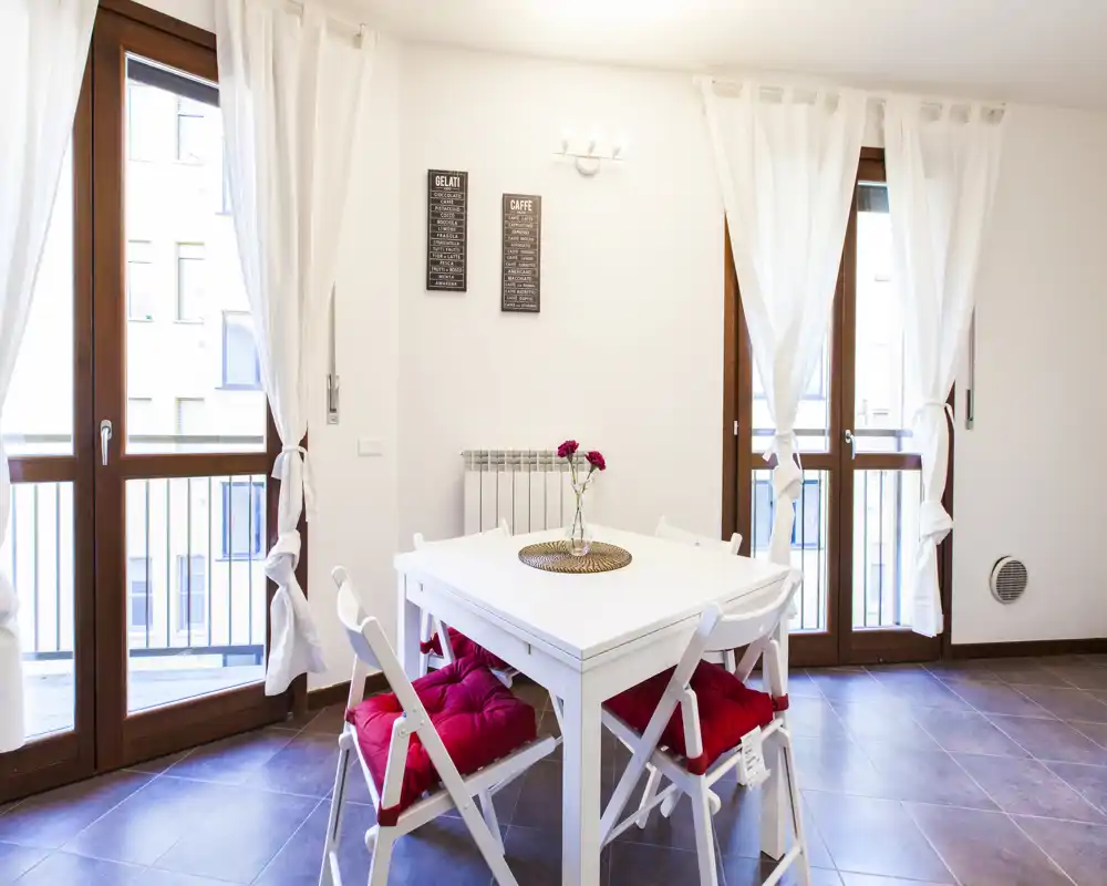 Zona pranzo luminosa e moderna con tavolo quadrato bianco e quattro sedie con cuscini rossi, ampie finestre e tendine leggere che lasciano entrare molta luce naturale.