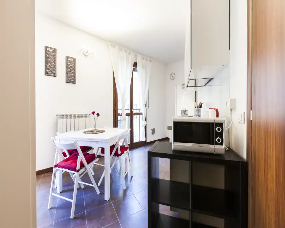 Zona pranzo luminosa e compatta con tavolo bianco, sedie pieghevoli con cuscini rossi e porta finestra che illumina l’ambiente — perfetta per annunci di monolocali o bilocali.