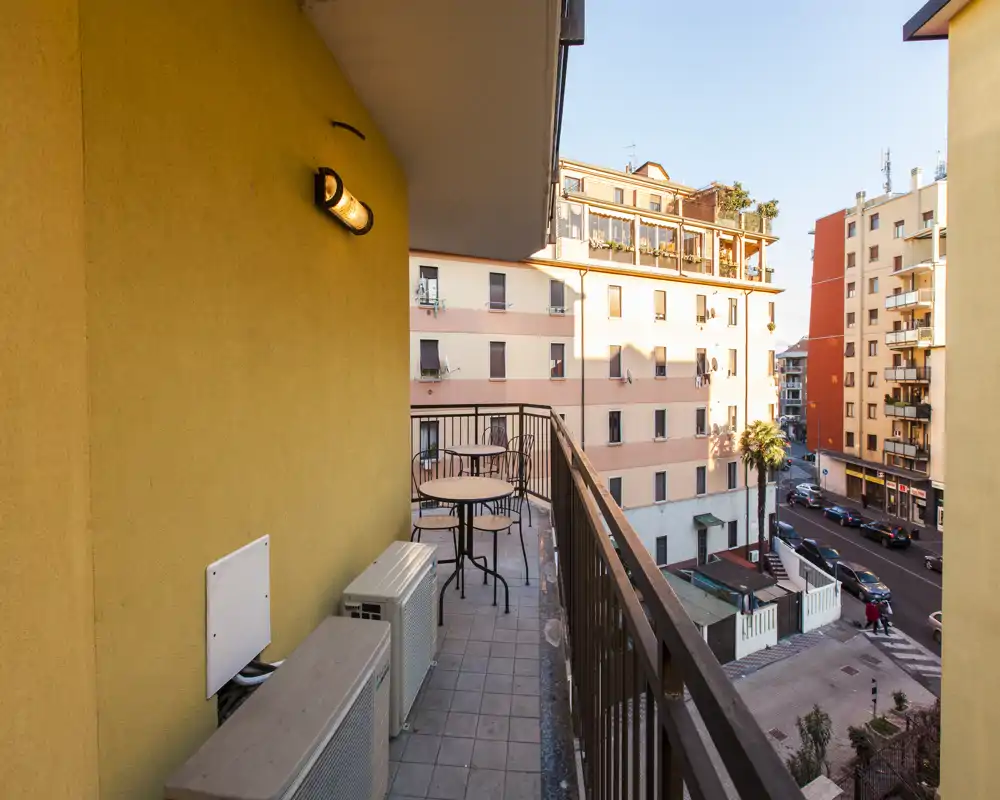 Balcone privato soleggiato con tavolo e sedie che si affaccia su una tranquilla via cittadina — ideale per colazioni o relax serali.