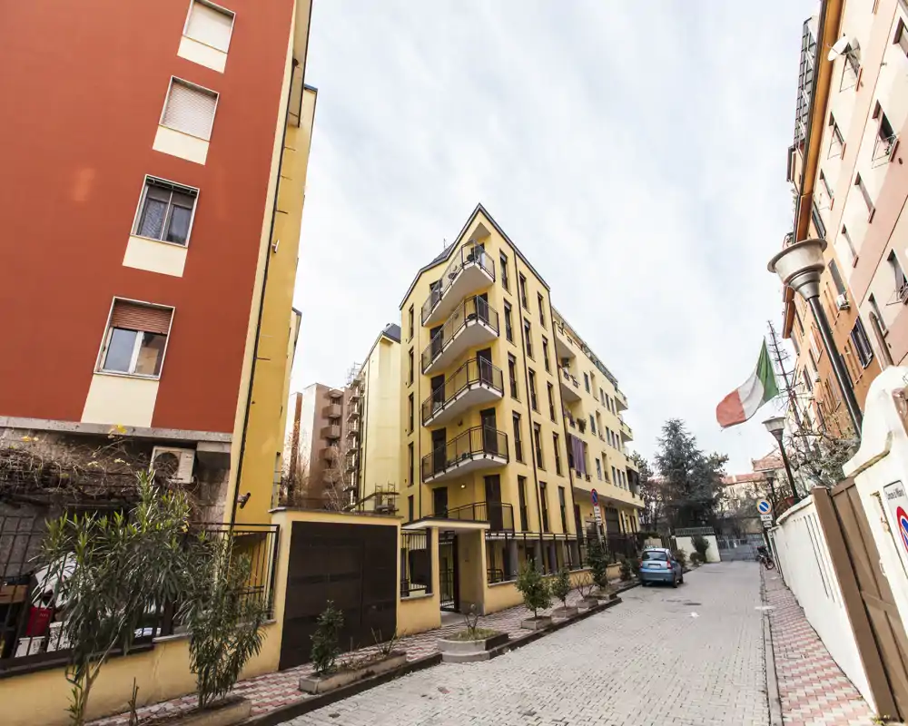 Vista esterna di una via residenziale con un palazzo giallo, piccoli vasi piante e auto parcheggiate; illuminazione chiara e composizione grandangolare.