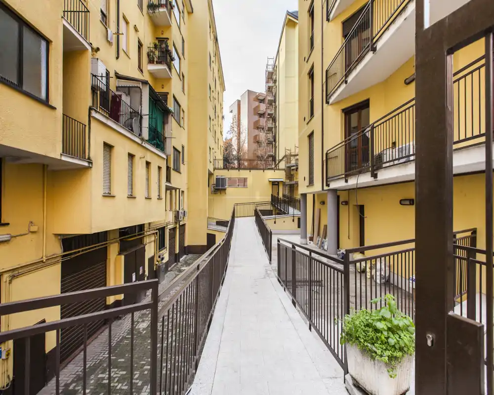 Cortile esterno condominiale con passerella pedonale sopraelevata tra palazzine gialle, ringhiere in metallo e vasi con piante.