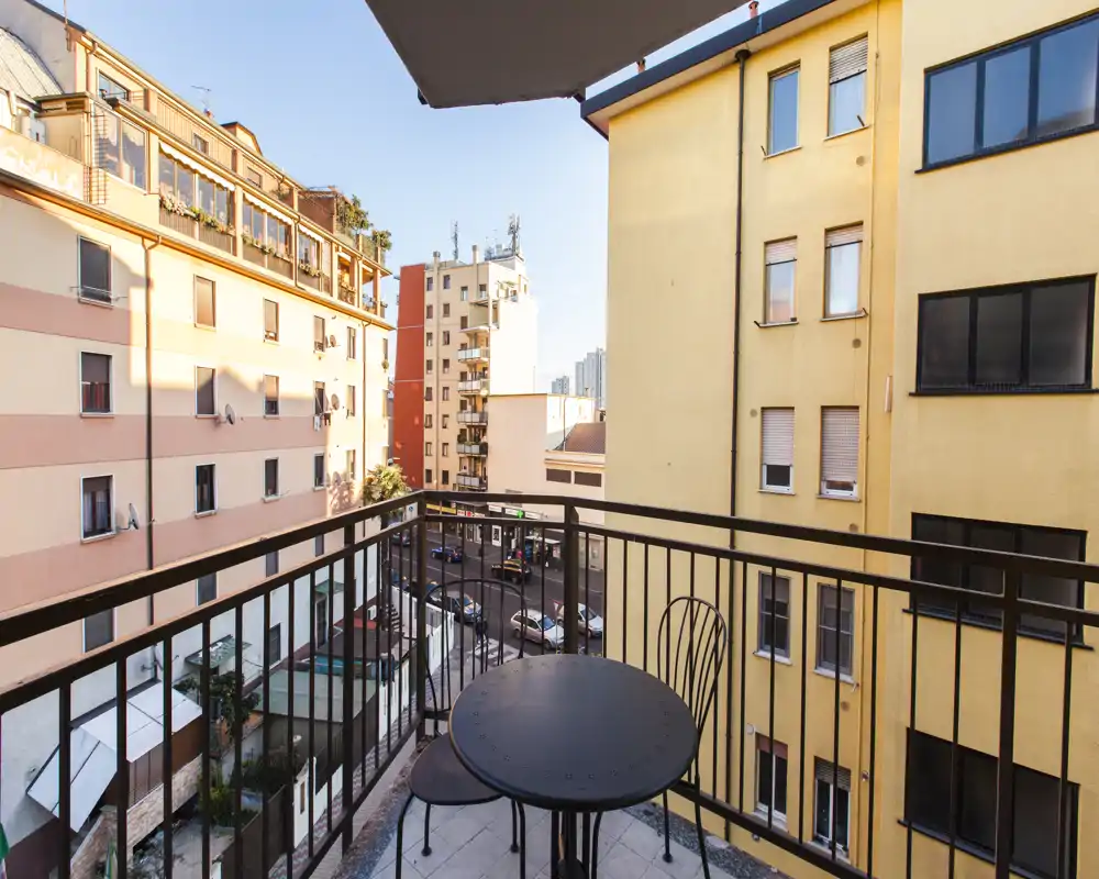 Piccolo balcone soleggiato con tavolino tondo in metallo e due sedie, affacciato su edifici residenziali e una strada tranquilla — perfetto per il caffè mattutino.