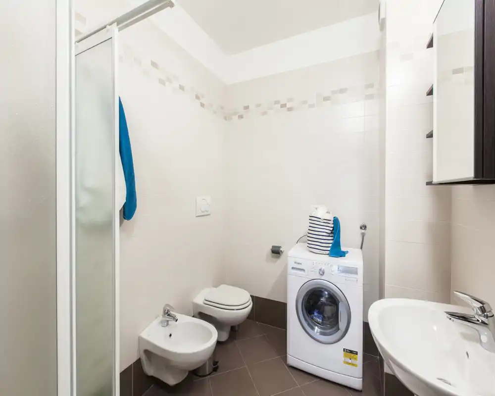 Bagno moderno e luminoso con doccia, bidet, wc sospeso, lavabo e lavatrice; piastrelle neutre e arredo semplice, perfetto per annunci di affitto.