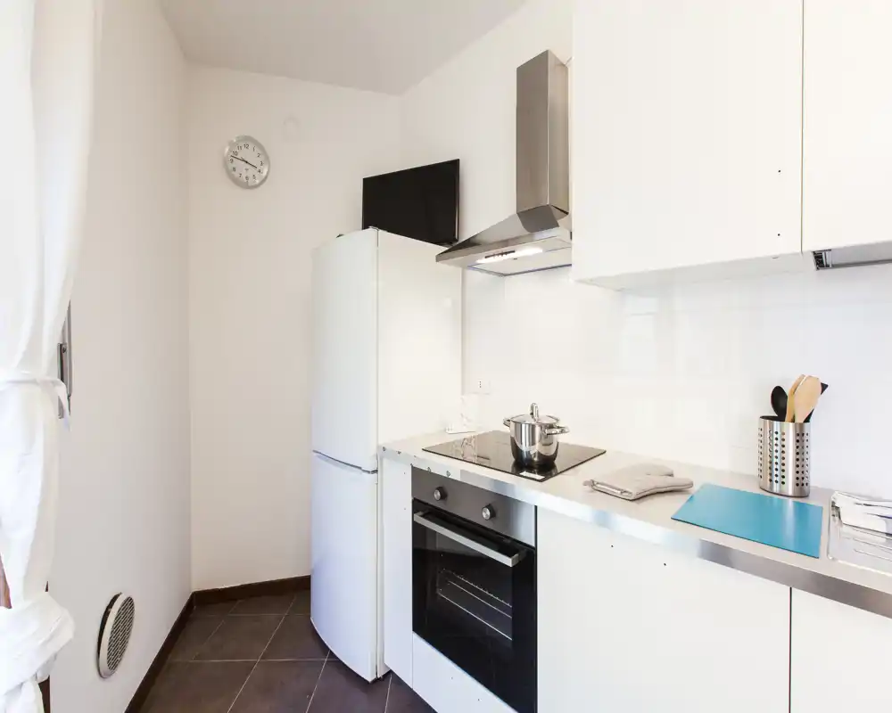Cucina moderna compatta con mobili bianchi, piano a induzione, forno incassato e frigorifero; ambiente pulito e luminoso perfetto per un appartamento piccolo.