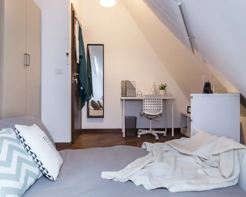 Dormitorio en el ático, acogedor, con una cama amplia en primer plano, tejidos suaves y un pequeño escritorio bajo el techo inclinado. Tonos neutros, suelo de madera e iluminación equilibrada que aportan calidez.