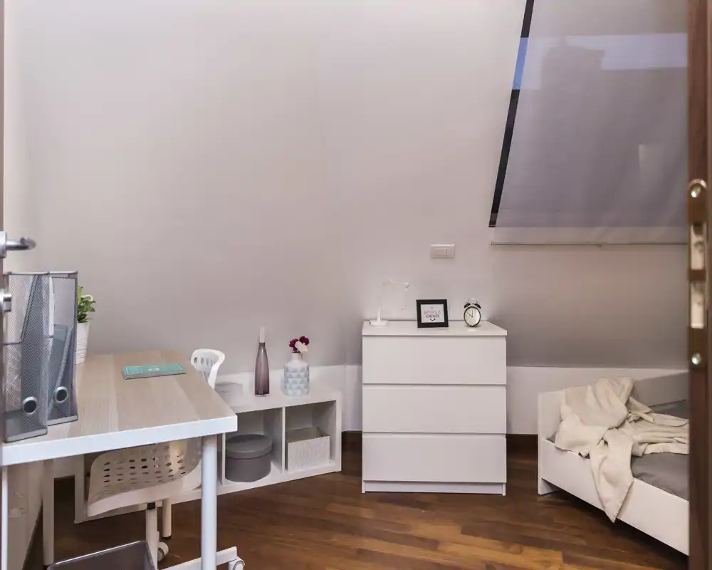 Rincón de dormitorio abuhardillado acogedor con una cama pequeña, cómoda blanca y un escritorio ordenado: luminoso, minimalista y bien decorado para un espacio compacto.