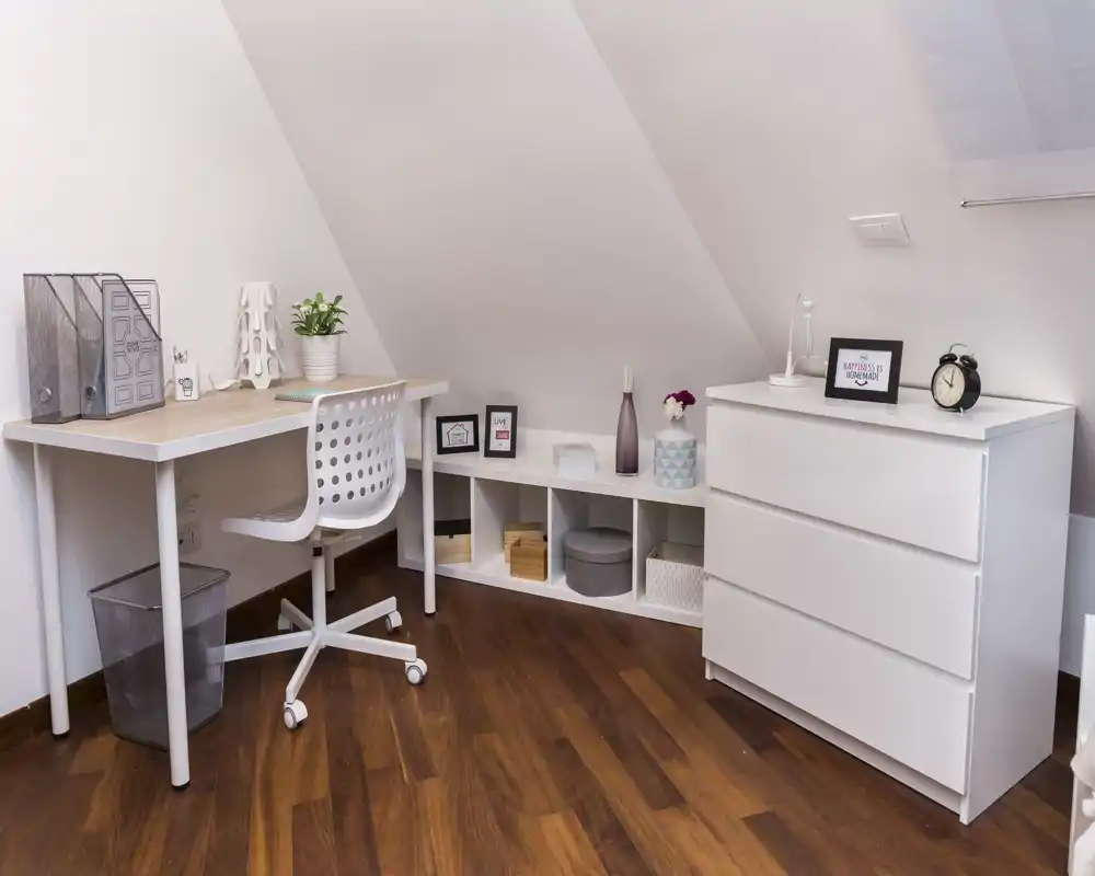 Espacio de trabajo en buhardilla luminoso y ordenado con escritorio blanco, silla ergonómica y almacenaje bajo—ideal para teletrabajo o estudio.