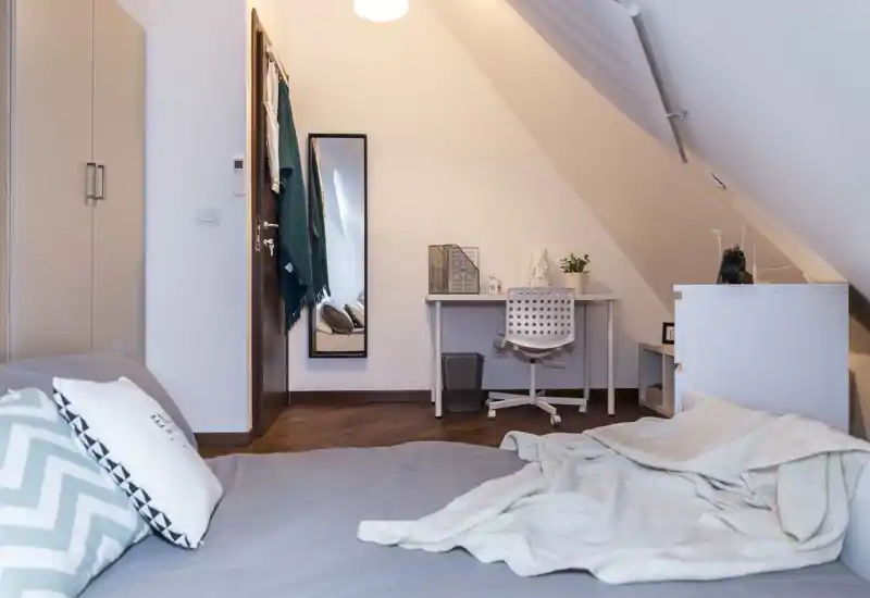 Dormitorio en el ático, acogedor, con una cama amplia en primer plano, tejidos suaves y un pequeño escritorio bajo el techo inclinado. Tonos neutros, suelo de madera e iluminación equilibrada que aportan calidez.