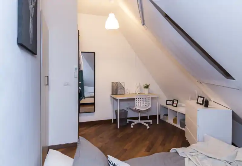 Dormitorio en buhardilla luminoso con cama acogedora en primer plano, pequeña mesa y silla bajo el techo inclinado, suelos de madera y decoración moderna sencilla.
