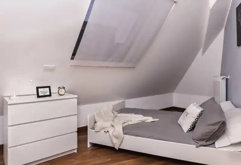 Dormitorio en ático luminoso con claraboya, cama minimalista blanca, ropa de cama gris y manta acogedora; suelo de madera cálido y cómoda sencilla para un aspecto moderno y limpio.