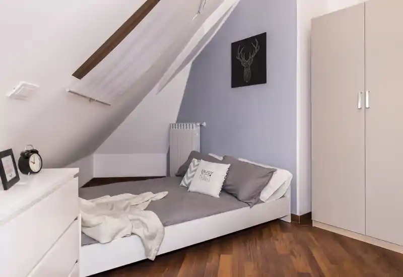 Pequeña habitación abuhardillada con cama individual, ropa de cama gris, cojines decorativos y suelos de madera cálidos; acogedora, de estilo sencillo y bien iluminada.
