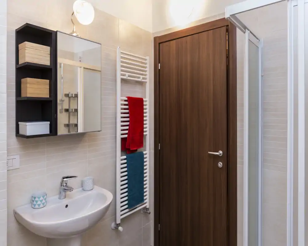 Baño limpio y bien iluminado con lavabo mural, armario con espejo, toallero radiador y cabina de ducha visible en el borde del encuadre.