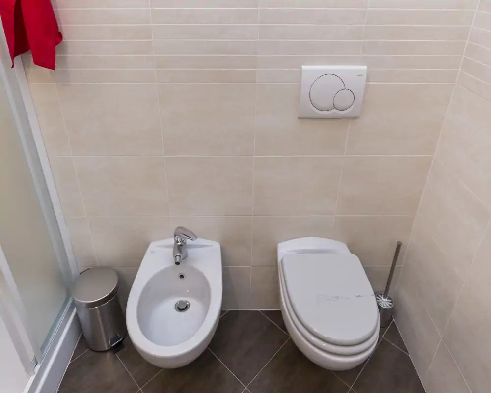 Baño compacto y moderno con inodoro, bidé, azulejos en paredes y suelo y la puerta de la ducha en el borde del encuadre.