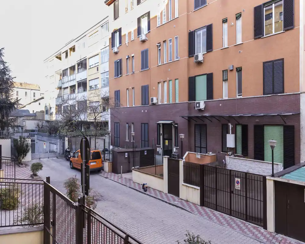 Patio residencial tranquilo con entrada privada, plantas en maceta y vehículo aparcado; apto para mostrar el área exterior común y el acceso.