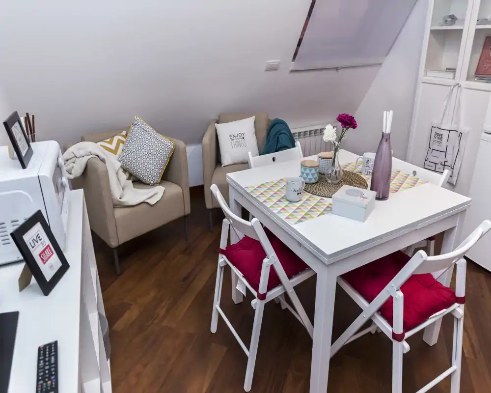 Rincón de salón/comedor luminoso y acogedor con mesa blanca, cuatro sillas con cojines rojos y dos butacas beige bajo techo inclinado — ideal para apartamentos pequeños.