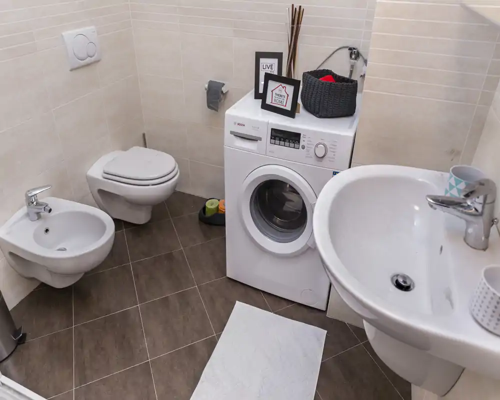 Baño moderno y limpio con inodoro suspendido, bidé, lavabo y lavadora de carga frontal integrada en el espacio. Baldosas neutras y distribución compacta, práctico para un piso.