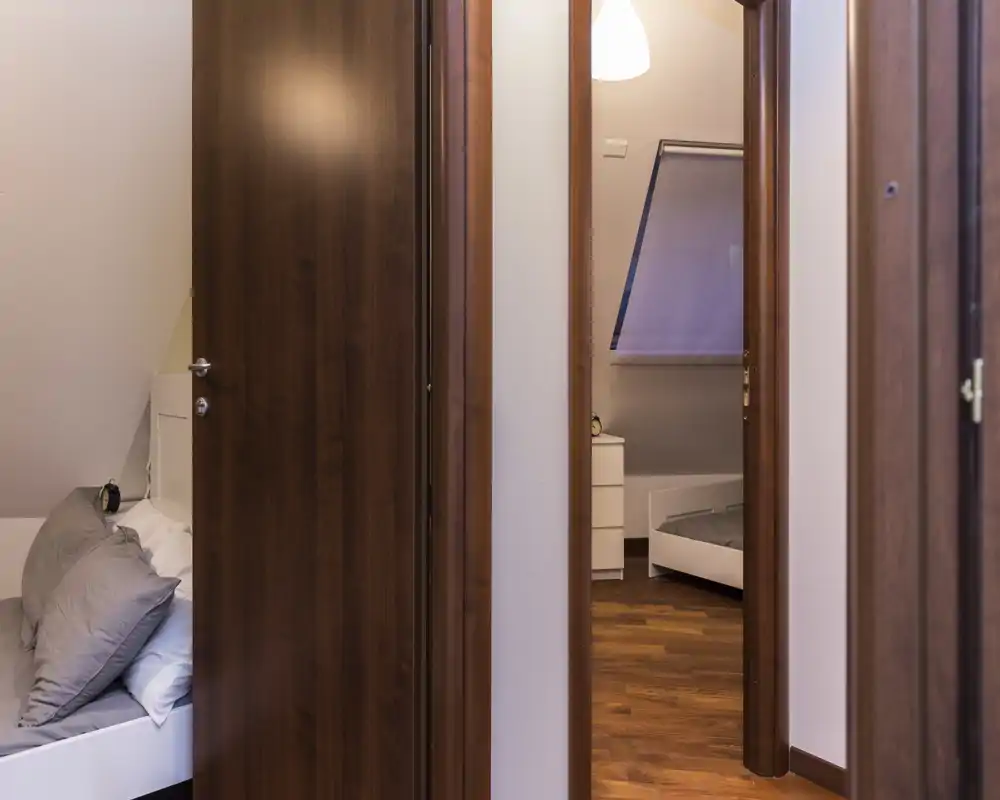Acogedora habitación abuhardillada con suelo de madera y ropa de cama suave; la foto muestra una cama parcialmente visible en primer plano y otra cama visible a través de la puerta, destacando las zonas de descanso y las mesillas.