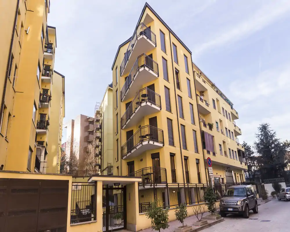 Vista exterior luminosa de un edificio residencial amarillo de varias plantas con balcones frente a una calle tranquila; se ven coches aparcados y una entrada con reja.