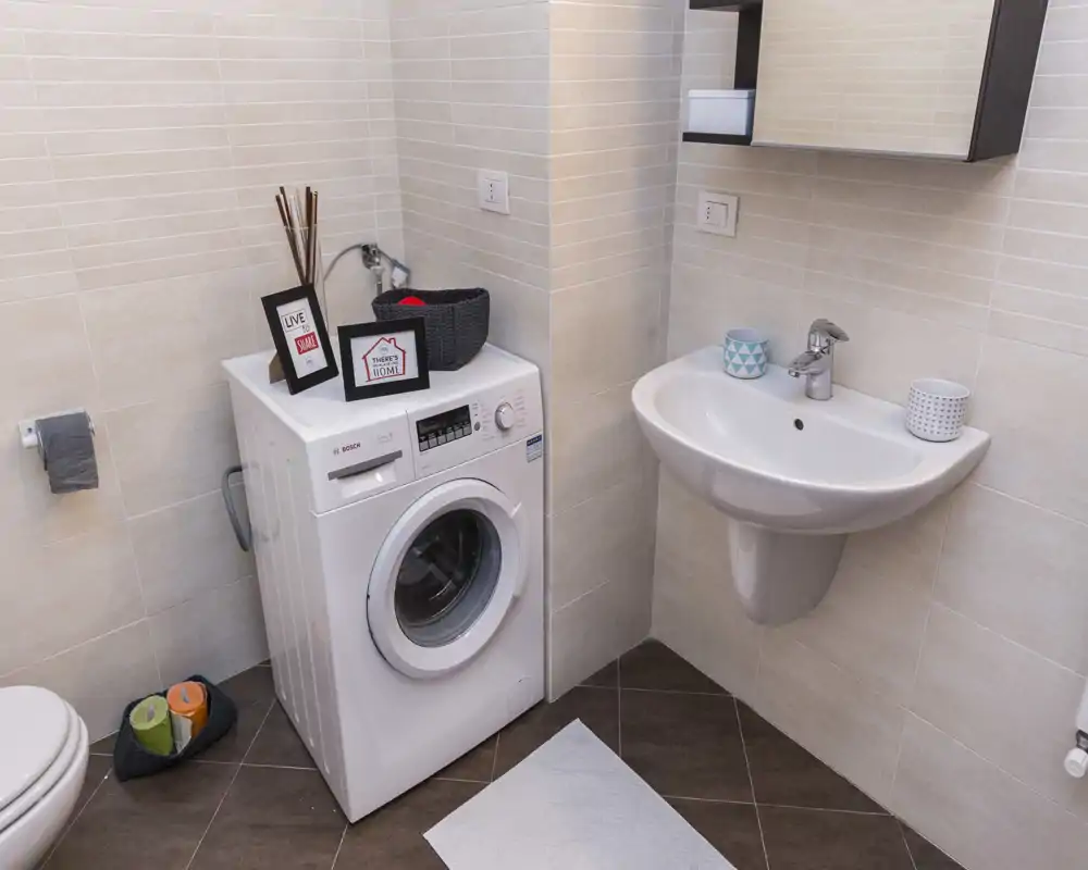 Baño compacto con lavabo suspendido, lavadora de carga frontal y azulejos beige neutro; espacio ordenado, bien iluminado y funcional, ideal para pisos pequeños.