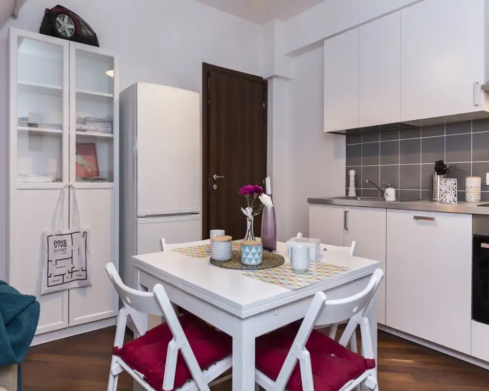 Cocina pequeña y moderna, luminosa, con mesa blanca, cuatro sillas con cojines rojos y mobiliario de cocina compacto con azulejos grises—perfecta para un piso pequeño.