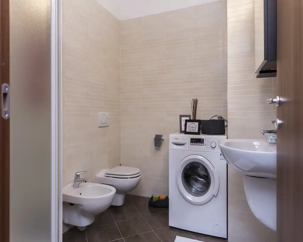 Baño moderno y compacto con inodoro y bidé suspendidos, lavadora de carga frontal y lavabo mural; azulejos neutros y presentación ordenada que ofrecen un espacio funcional.