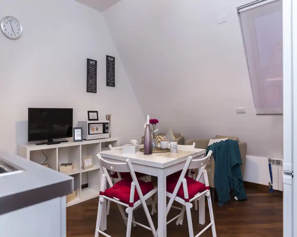 Acogedor rincón salón/comedor bien decorado con mesa blanca, sillas con cojines rojos, mueble TV y pequeño sofá con mantas — ideal para pisos compactos.