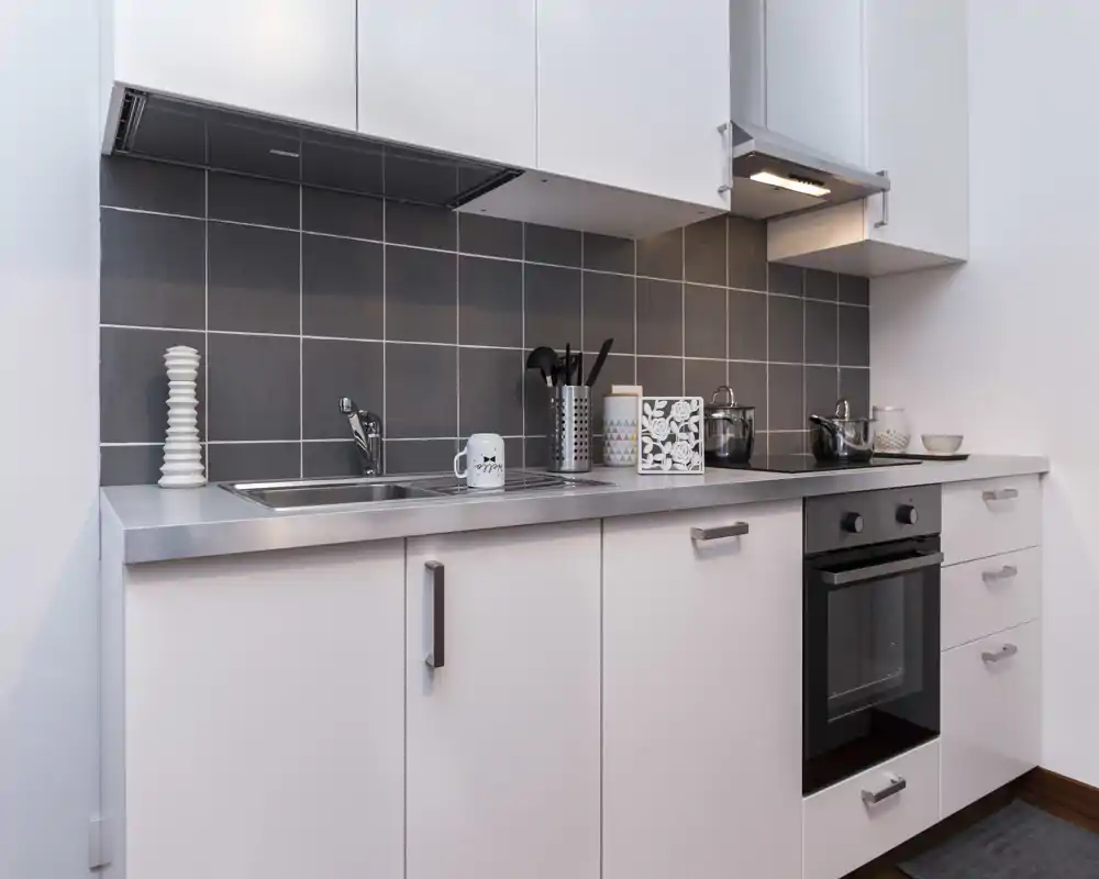 Cocina moderna y compacta con encimera de acero inoxidable, horno empotrado y fregadero; puesta en escena ordenada con utensilios y pequeños objetos decorativos.