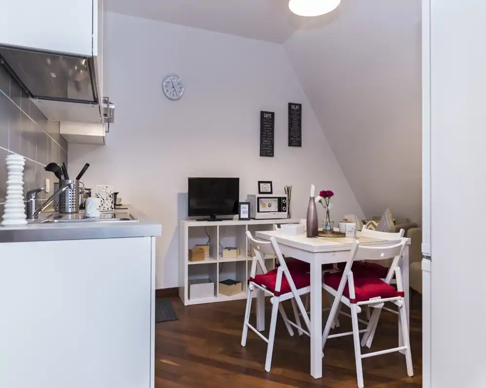 Zona de estar luminosa y acogedora con una pequeña mesa, estantería para TV y una cocina compacta—ideal para un estudio cómodo.