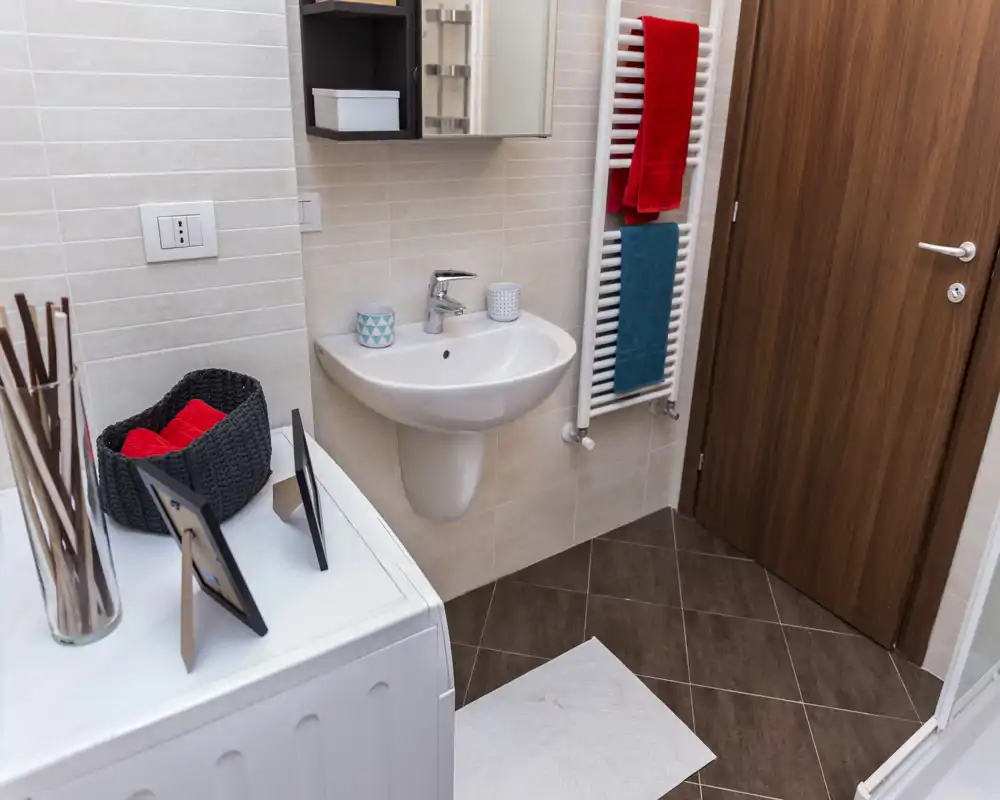 Baño compacto y bien iluminado con lavabo mural, toallero calefactado con toallas de colores, puerta efecto madera y suelo de baldosa; ordenado y moderno.
