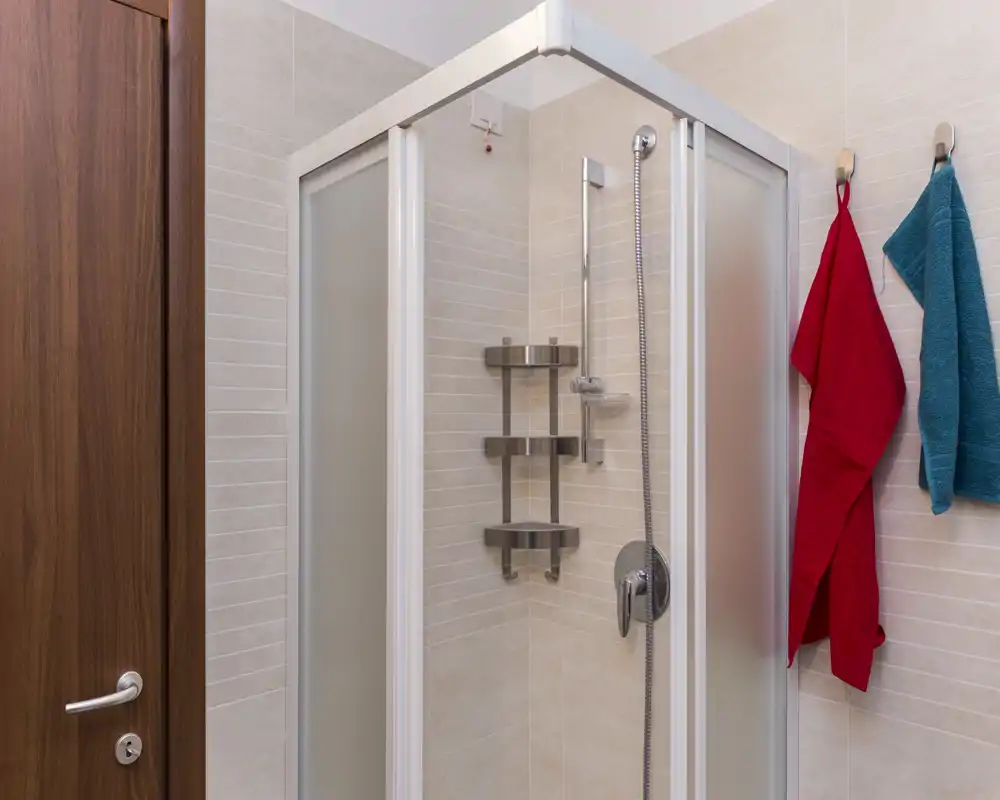Baño moderno y compacto con cabina de ducha en esquina, grifería cromada y paredes alicatadas; dos toallas de colores colgadas en ganchos.
